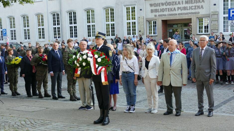 Strażniczka miejska niesie wieniec, za nią oficjalna delegacja.