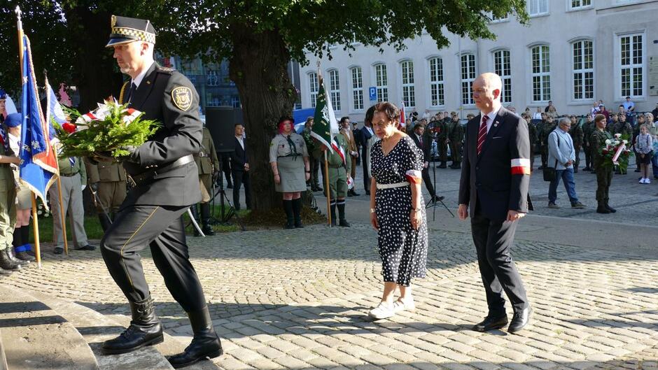 Strażnik miejski niesie wieniec, za nim dwuosobowa delegacja.