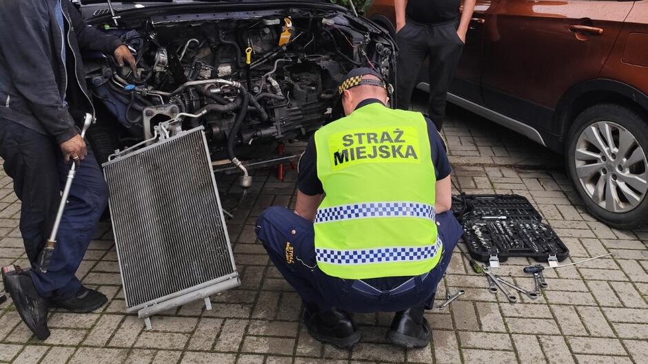 strażnik kuca przy samochodzie obok stoi mężczyzna z metalową rurką