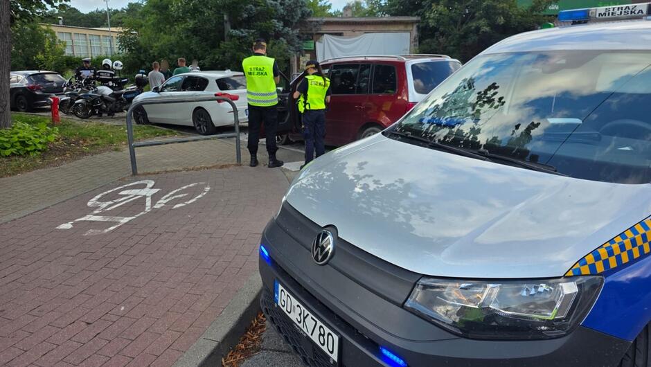 przód radiowozu straży miejskiej w tle policjanci na motocyklach zaparkowane na jezdni auta obok których stoją ludzie