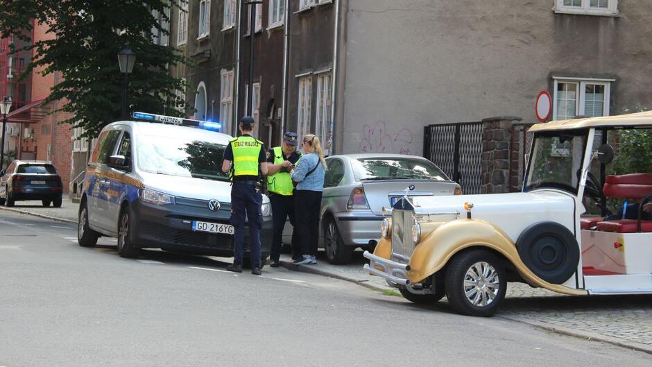 Strażnicy stoją obok radiowozu i meleksa i wypisują mandat. Obok nich stoi kobieta.