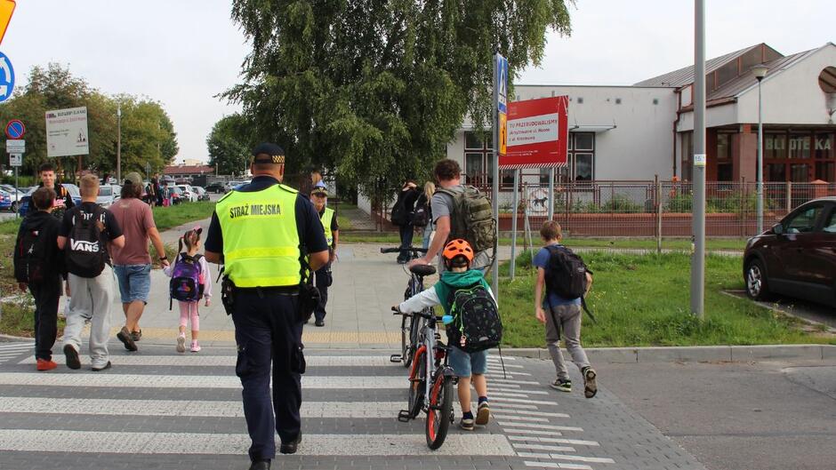 Strażnik miejski eskortuje przez przejście dla pieszych dwoje rowerzystów.