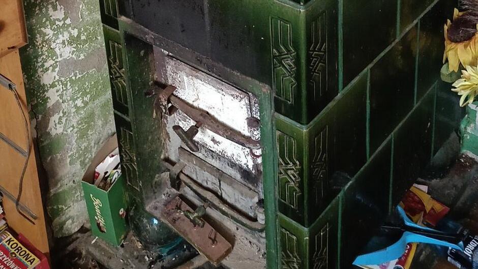 Zabytkowy piec kaflowy w zielonym kolorze z metalowymi drzwiczkami i zamkiem, noszący ślady użytkowania i osmalenia. Kafle zdobią dekoracyjne wzory. Po lewej stronie znajduje się drewniana półka z różnymi przedmiotami, w tym pudełkiem z napisem 'KORONA'. Po prawej stronie widoczne są rozrzucone materiały opakowaniowe oraz prawdopodobnie kwiaty. Tło stanowi ściana z łuszczącą się farbą w odcieniach zieleni i bieli.