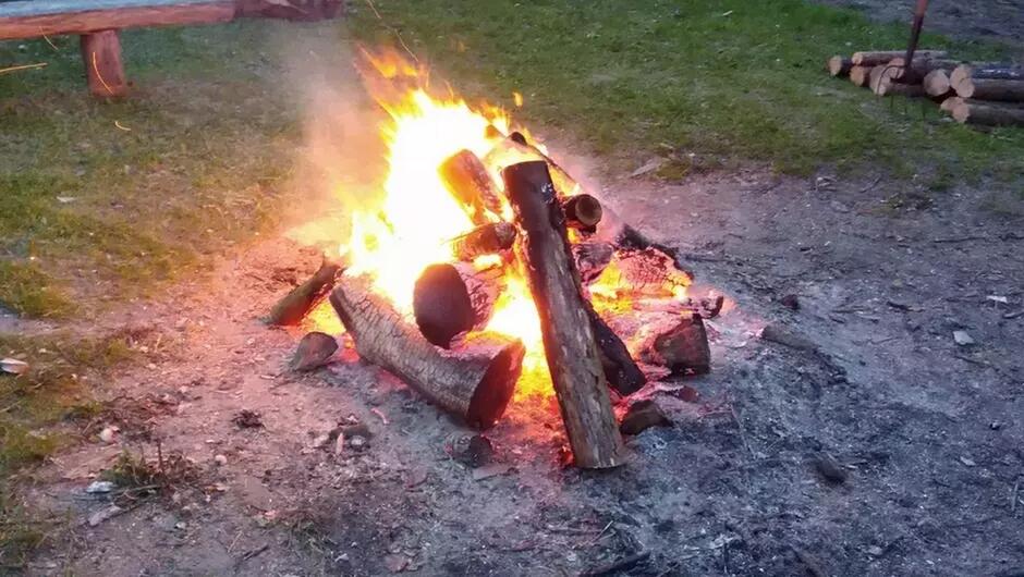 Na zdjęciu widoczne jest rozpalone ognisko z kilkoma jasnymi, płonącymi polanami. Ognisko znajduje się na skrawku gołej ziemi, otoczonym trawą. W tle po lewej stronie znajduje się drewniana ławka, a po prawej stos pociętych drewien. Płomienie są wysokie, a z ognia unosi się dym.