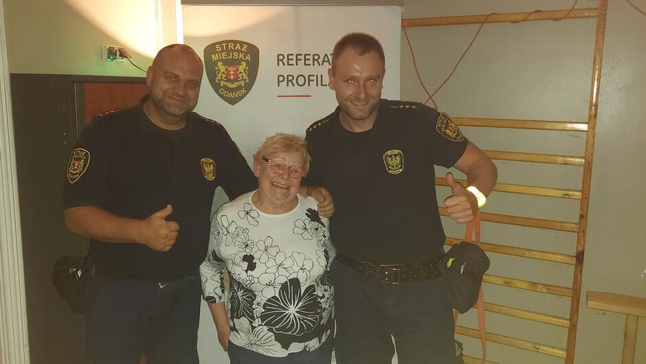 dwóch strażników pozuje do zdjęcia z seniorką stoją na tle baneru Referat Profilaktyki