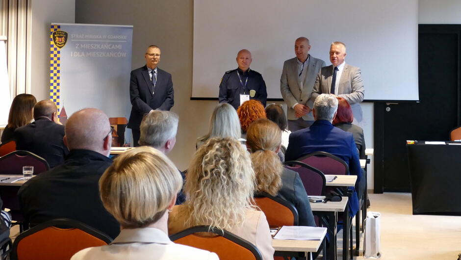 komendant Leszek Walczak otwiera konferencję stojąc przed siedzącymi na krzesłach uczestnikami, obok Patryk Punsewicz główny księgowy, Paweł Jankowski naczelnik wydziału logistyki, Marek Sawicki kierownik Referatu Kadr