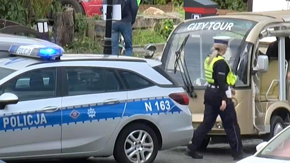 Zdjęcie przedstawia scenę miejską z udziałem służb porządkowych. Na pierwszym planie widoczny jest radiowóz policyjny z oznaczeniem „POLICJA” oraz numerem identyfikacyjnym „N 163”. W jego kierunku zmierza funkcjonariusz w mundurze policyjnym. Obok znajduje się pojazd turystyczny z napisem „CITY TOUR” na przodzie, który przypomina elektryczny wózek wykorzystywany do przewozu turystów. W tle widoczne są inne osoby — jedna stoi przy czerwonym samochodzie, a druga znajduje się wewnątrz pojazdu turystycznego. Otoczenie wskazuje na teren miejski z obecnością zieleni i budynków w tle.