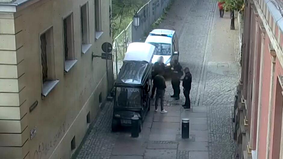 Zdjęcie przedstawia wąską, brukowaną uliczkę otoczoną budynkami po obu stronach. Po lewej stronie zaparkowany jest niewielki pojazd z napisem „CITY TOUR”, sugerujący, że może to być pojazd wykorzystywany do zwiedzania miasta. Nieco dalej, w głębi ulicy, znajduje się kolejny pojazd — radiowóz straży miejskiej. Przy pojeździe „CITY TOUR” stoi grupa osób, które wydają się być zaangażowane w rozmowę lub jakąś czynność. Scena ma miejsce w ciągu dnia. Budynki widoczne na zdjęciu mają liczne okna, a na ścianie jednego z nich (po lewej stronie) można dostrzec graffiti.