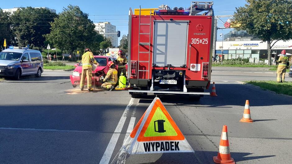 Na zdjęciu przedstawiona jest scena wypadku drogowego: Czerwony samochód osobowy uległ kolizji — widoczne są uszkodzenia.
Na miejscu zdarzenia pracują strażacy w żółtych mundurach, którzy zabezpieczają teren i udzielają pomocy.
Obecny jest wóz strażacki oznaczony numerem 305/25, zaparkowany tuż obok miejsca zdarzenia.
Ustawiono trójkątny znak ostrzegawczy z wykrzyknikiem i napisem WYPADEK , informujący o zagrożeniu.
Teren jest zabezpieczony pachołkami drogowymi, które kierują ruchem.
W tle widoczne są inne pojazdy, drzewa, budynki oraz znak przejścia dla pieszych.