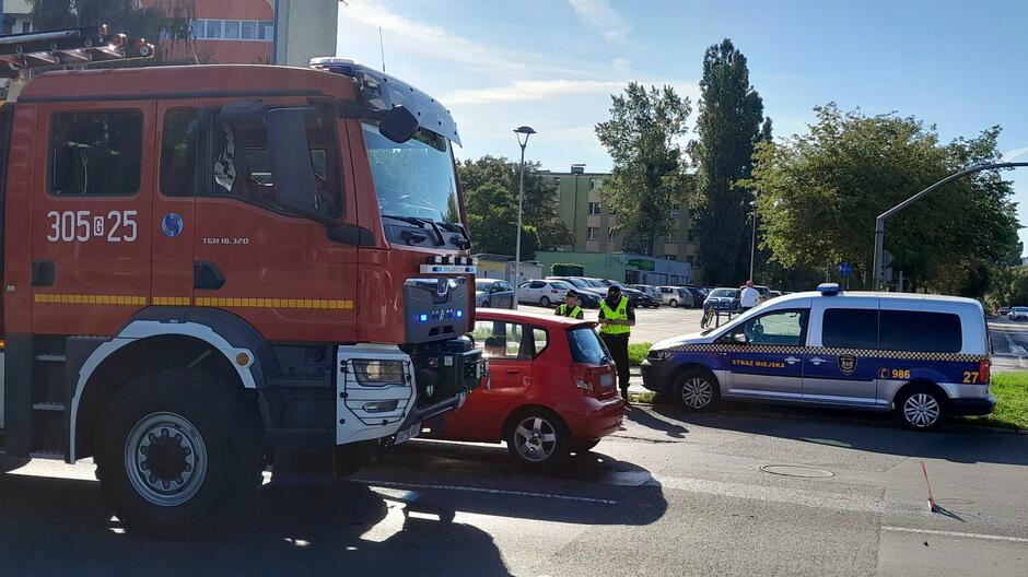 Na zdjęciu widoczna jest scena kolizji drogowej z udziałem czerwonego samochodu osobowego, wozu strażackiego oraz radiowozu Straży Miejskiej.
Szczegóły obrazu: Czerwony samochód ma uszkodzony przód, co wskazuje na miejsce uderzenia.
Wóz strażacki oznaczony numerem 305[G]25 ma włączone światła ostrzegawcze.
Radiowóz Straży Miejskiej oznaczony jest numerem 986.
Trzech funkcjonariuszy w kamizelkach odblaskowych znajduje się w pobliżu pojazdów — prawdopodobnie oceniają sytuację lub kierują ruchem.
W tle widoczne są drzewa oraz budynki mieszkalne, a niebo jest częściowo zachmurzone.