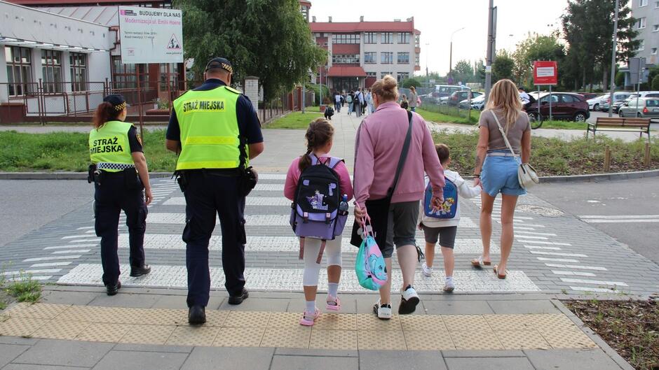 Dwóch funkcjonariuszy Straży Miejskiej w kamizelkach odblaskowych pomaga grupie dorosłych i dzieci przejść przez przejście dla pieszych. Dzieci mają plecaki, co sugeruje, że sytuacja ma miejsce w pobliżu szkoły lub przedszkola. W tle widoczny jest budynek edukacyjny.