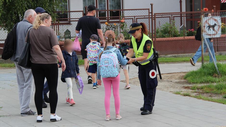 Funkcjonariusz Straży Miejskiej rozmawia z dzieckiem niosącym plecak. W tle znajdują się inni dorośli i dzieci, co wskazuje na szkolne otoczenie. Sytuacja wygląda na przyjazną i edukacyjną – możliwe, że funkcjonariusz udziela wskazówek dotyczących bezpieczeństwa.