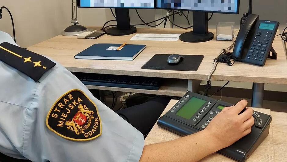 Zdjęcie przedstawia pracownika Straży Miejskiej w Gdańsku, siedzącego przy stanowisku monitoringu. Ubrany jest w mundur z widocznym emblematem „STRAŻ MIEJSKA GDAŃSK” na rękawie. Na biurku znajdują się dwa monitory, telefon, notatnik z pomarańczowym długopisem, oraz urządzenia elektroniczne, w tym panel komunikacyjny, na którym spoczywa ręka funkcjonariusza. Scena ukazuje pracę w centrum monitoringu miejskiego.