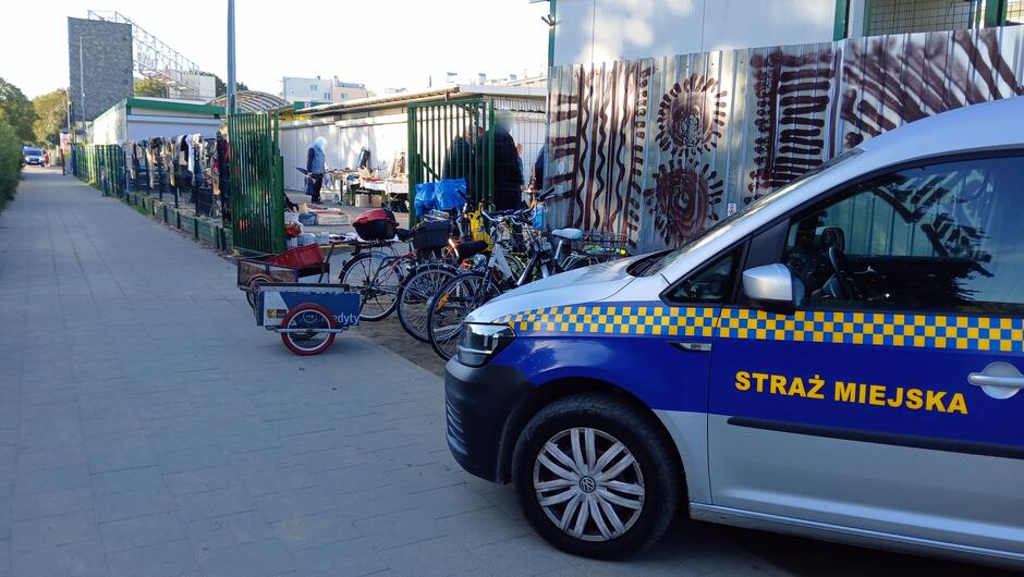 Scena z miejskiego targowiska w Gdańsku. Po prawej stronie widoczny jest zaparkowany radiowóz Straży Miejskiej z charakterystycznym niebiesko-białym oznakowaniem i żółtą szachownicą. Po lewej stronie, za zielonym ogrodzeniem, znajdują się stoiska handlowe z różnorodnym asortymentem. Przy ogrodzeniu zaparkowano kilka rowerów, w tym jeden z przyczepką. W tle widoczne są budynki i drzewa, co podkreśla miejski charakter miejsca. Obecność służb porządkowych wskazuje na działania mające na celu utrzymanie ładu w okolicy targowiska.