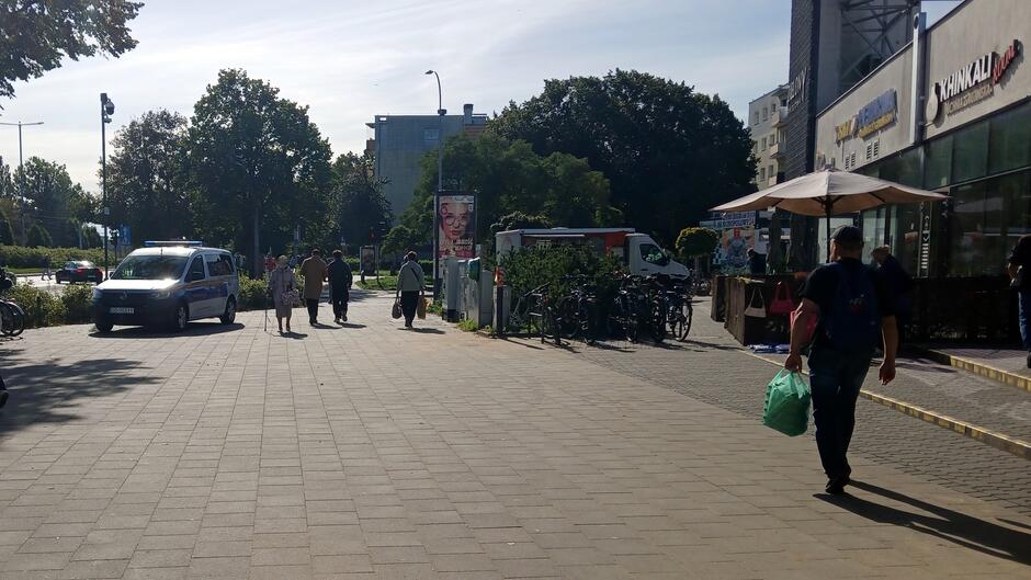 Ulica w miejskiej przestrzeni w słoneczny dzień. Na chodniku spacerują przechodnie, niektórzy z torbami zakupowymi. Po lewej stronie widoczny jest zaparkowany samochód oraz drzewa wzdłuż ulicy. Po prawej stronie znajduje się budynek z szyldami lokali gastronomicznych, w tym „Khinkali”, oraz ogródek z parasolami. W pobliżu zaparkowano również rowery. W tle widać kolejne budynki i zieleń.