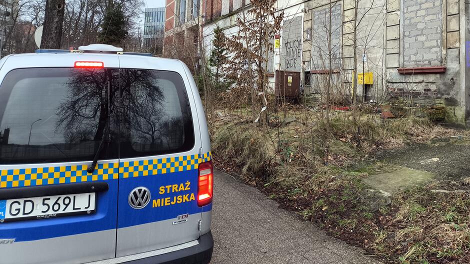 Na fotografii widoczny jest tylny fragment pojazdu Straży Miejskiej o numerze rejestracyjnym GD 569LJ, zaparkowany na wąskiej drodze. Pojazd stoi obok zaniedbanego budynku, którego okna i drzwi są zamknięte. Otoczenie budynku porasta dzika roślinność, a na jego ścianach znajdują się graffiti oraz tabliczki ostrzegawcze. Sceneria sugeruje interwencję lub asystę funkcjonariuszy w miejscu wymagającym działań porządkowych.
