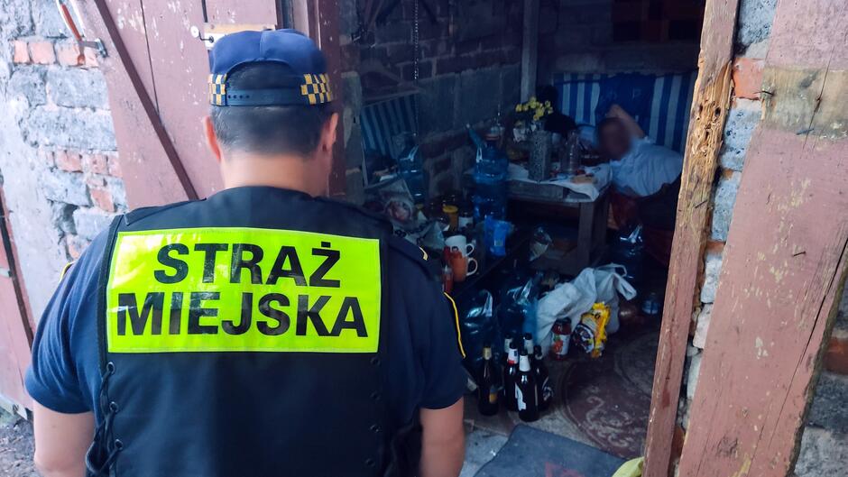 Na fotografii widoczny jest funkcjonariusz Straży Miejskiej, stojący tyłem — na jego mundurze znajduje się napis „STRAŻ MIEJSKA”. Strażnik znajduje się przy wejściu do niewielkiego, zagraconego pomieszczenia lub altany. W środku leży osoba na prowizorycznym łóżku lub kanapie. Wnętrze jest pełne różnych przedmiotów: butelek, toreb i domowych rzeczy porozrzucanych wokół. Scena przedstawia prawdopodobnie interwencję lub asystę strażnika wobec osoby przebywającej w trudnych warunkach.