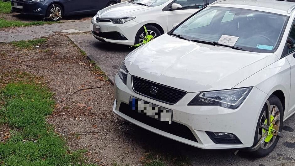 Na fotografii widoczne są dwa białe samochody zaparkowane na parkingu, oba z założonymi jaskrawozielonymi blokadami na koła. Na przedniej szybie jednego z pojazdów znajduje się kartka papieru, prawdopodobnie informująca o nałożonej blokadzie lub zawierająca mandat. W tle widać trzeci, ciemniejszy samochód zaparkowany przy krawężniku oraz fragment chodnika i trawnika.