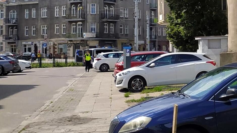 Na fotografii widoczna jest uliczna scena w miejskiej zabudowie. Po obu stronach ulicy zaparkowanych jest kilka samochodów — m.in. białe, czerwone i niebieskie. W tle znajdują się wielopiętrowe budynki mieszkalne z balkonami. W centralnej części zdjęcia stoi osoba w kamizelce odblaskowej, prawdopodobnie funkcjonariusz Straży Miejskiej, który nadzoruje sytuację lub kontroluje parkowanie. Obok widoczny jest parkomat, a całość przedstawia typowy fragment miejskiej infrastruktury.