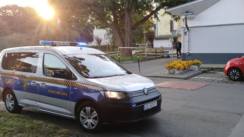 Straż Miejska podczas kontroli w rejonie Zielonego Rynku. Na zdjęciu widoczny radiowóz z włączonymi światłami ostrzegawczymi zaparkowany przy ulicy Czerwony Dwór. W tle ustawione donice z kwiatami oraz przechodzień zmierzający w kierunku budynku. Ujęcie wykonane o zachodzie słońca, co nadaje scenie ciepły klimat.