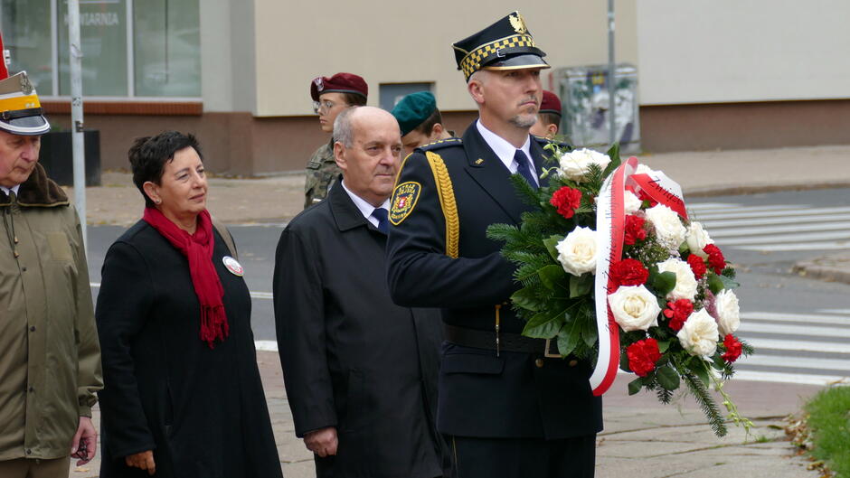 Zdjęcie przedstawia moment uroczystości upamiętniającej, odbywającej się w przestrzeni miejskiej – prawdopodobnie przy skrzyżowaniu ulic. W centrum kadru znajduje się umundurowany funkcjonariusz trzymający dużą wiązankę z czerwonych i białych kwiatów, ozdobioną zielenią i wstążką. Obok niego stoją inni uczestnicy wydarzenia – część w mundurach wojskowych, część w ubraniach cywilnych. W tle widoczne są budynki, co wskazuje na miejskie otoczenie.