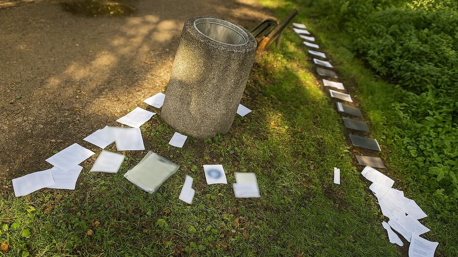 Na zdjęciu widać porzucone dokumenty leżące na trawie w pobliżu ulicznego kosza. Część z nich została rozrzucona wzdłuż ścieżki i przy ławce. Widoczne papiery zawierały dane osobowe, które mogły trafić w niepowołane ręce. Dzięki reakcji spacerowiczki i interwencji strażników miejskich udało się odnaleźć właściciela i zapobiec potencjalnemu zagrożeniu.