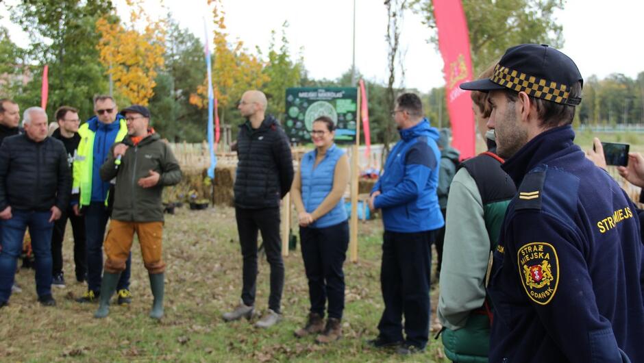 Kilka osób stoi w rzędzie podczas krótkiego przemówienia lub instruktażu. W tle widoczne są banery informacyjne i elementy przygotowanego terenu do sadzenia. Na pierwszym planie znajduje się funkcjonariusz Straży Miejskiej w umundurowaniu.