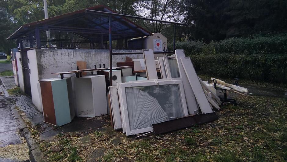 Na zdjęciu widać miejsce przeznaczone na składowanie odpadów wielkogabarytowych, prawdopodobnie przy altanie śmietnikowej. W centralnej części obrazu znajduje się duża sterta zdemontowanych elementów wyposażenia wnętrz – głównie drzwi, fronty szafek, blaty oraz ramy okienne z szybami. Widać również kilka korpusów mebli kuchennych i fragmenty stołów. Wszystko jest ułożone w stosy na chodniku, obok betonowej konstrukcji altany z zadaszeniem z niebieskiej blachy.
W tle po lewej stronie widoczna jest część ulicy, a po prawej – gęsty żywopłot i drzewa. Na ścianie altany znajduje się napis „1844” oraz symbol serca w czerwonym kolorze. Na ziemi leżą mokre liście, co sugeruje, że zdjęcie wykonano jesienią, po deszczu (widoczne mokre plamy na chodniku).