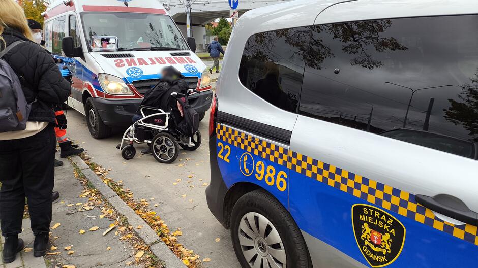 Na zdjęciu widoczna jest scena interwencji służb ratunkowych w miejskim otoczeniu. Ambulans stoi zaparkowany przy ulicy, z napisem „AMBULANS” widocznym w odbiciu lustrzanym na przedzie pojazdu.
Obok ambulansu znajduje się osoba na wózku inwalidzkim, której pomagają ratownicy medyczni.
Radiowóz Straży Miejskiej Gdańsk oznaczony numerami „22” i „986” oraz herbem jednostki stoi w pobliżu.
Na miejscu obecnych jest kilka osób, w tym jedna ubrana w czarną kurtkę i plecak.
W tle widoczne są drzewa oraz zabudowania, co wskazuje na miejskie otoczenie.