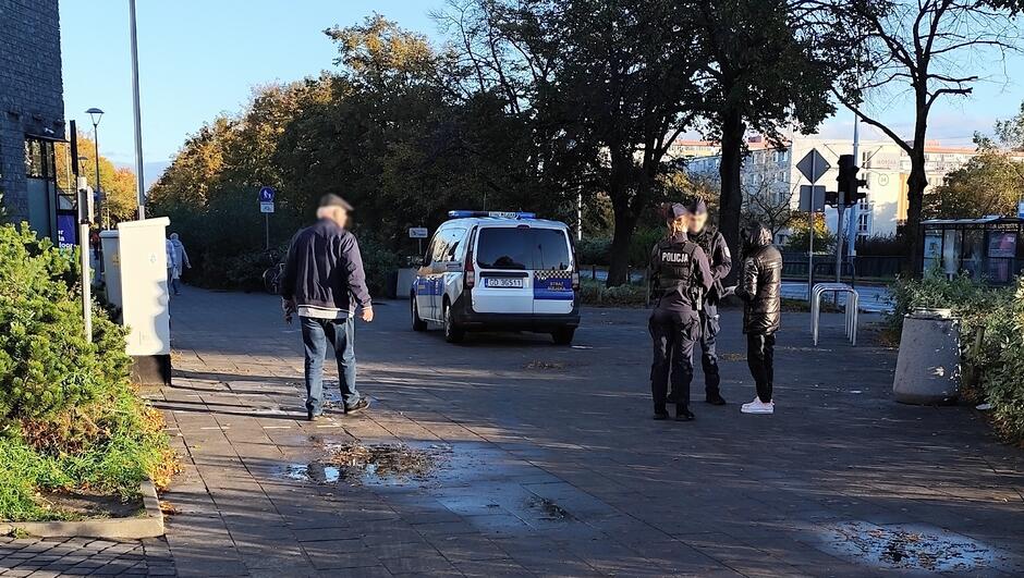Na zdjęciu widoczna jest scena z udziałem służb mundurowych w przestrzeni miejskiej: Na pierwszym planie stoi radiowóz policyjny zaparkowany na utwardzonym terenie.
Obok pojazdu znajdują się dwaj funkcjonariusze, którzy rozmawiają z osobą ubraną na ciemno.
W tle widać drzewa z jesiennym listowiem, budynek oraz znaki drogowe.
Po lewej stronie kadru ktoś oddala się od miejsca zdarzenia.
Na nawierzchni widoczne są kałuże, co sugeruje, że niedawno padał deszcz.
Długie cienie wskazują na porę dnia — prawdopodobnie wczesny poranek lub późne popołudnie.