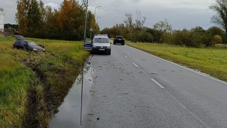 samochód opel corsa stoi na łące na zdrodze zaparkowany radiowóz z włączonymi światłami niebieskimi. obok niego przejeżdza czarny samochód terenowy.
