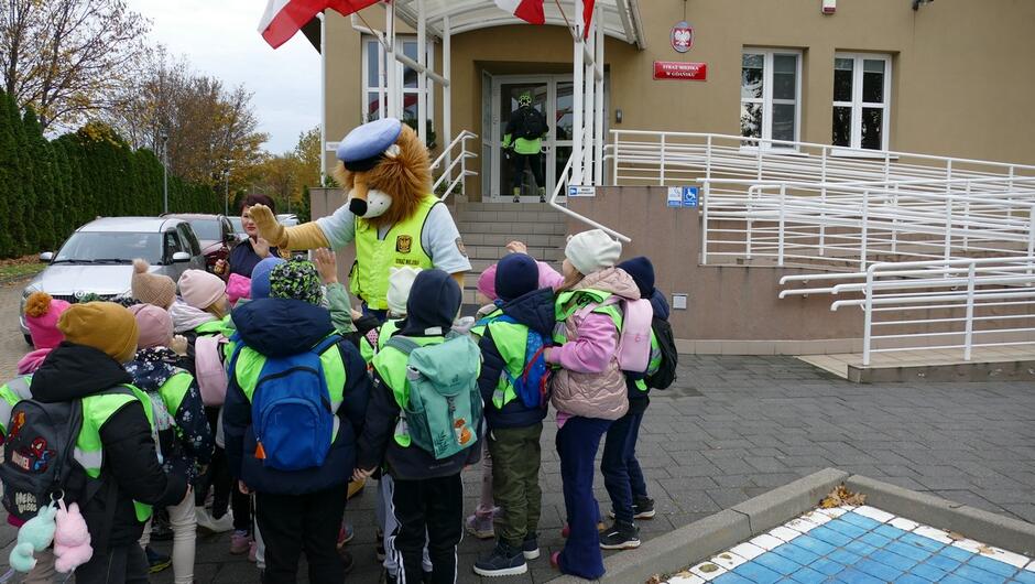 Grupa dzieci w odblaskowych kamizelkach stoi przed budynkiem urzędu lub instytucji publicznej, oznaczonym tablicą „Straż Miejska”. Nad wejściem widoczne są biało-czerwone flagi. Dzieci otaczają postać w stroju maskotki lwa ubranego w mundur straży miejskiej i kamizelkę odblaskową. W tle znajduje się samochód oraz osoba w mundurze przy drzwiach wejściowych.