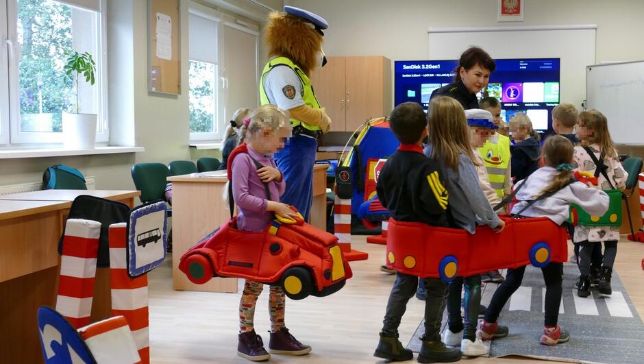 W sali edukacyjnej dzieci uczestniczą w zajęciach dotyczących bezpieczeństwa drogowego. Kilkoro z nich ma na sobie kostiumy w kształcie samochodów. W pomieszczeniu ustawione są makiety znaków drogowych i elementy imitujące ulicę. Obok dzieci stoi maskotka lwa w mundurze straży miejskiej.