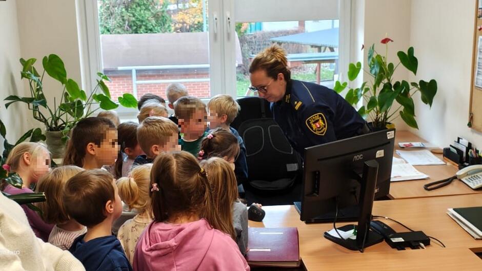 W biurze z biurkami, komputerem i roślinami grupa dzieci stoi przy stanowisku komputerowym. Obok nich znajduje się osoba w mundurze straży miejskiej oraz maskotka lwa. Wygląda na to, że dzieci poznają pracę straży miejskiej w praktyce.
