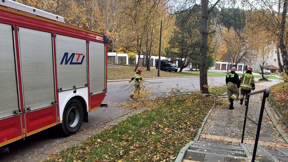 Zdjęcie przedstawia akcję strażaków na spokojnej, osiedlowej ulicy w jesiennej scenerii.
Po lewej stronie znajduje się czerwony wóz strażacki z widocznym oznaczeniem „MT”.
Na środku zdjęcia strażak w pełnym umundurowaniu i kasku usuwa z drogi duży, złamany konar drzewa.
Po prawej stronie dwóch strażaków oddala się w kierunku chodnika, prawdopodobnie po zakończeniu działań.
Widać drzewa z żółtymi i brązowymi liśćmi, co wskazuje na jesień.
W oddali stoją garaże i zaparkowane samochody.
Na chodniku widać barierki i schody prowadzące w dół.
Konar jest dość duży, z liśćmi, co sugeruje, że został oderwany przez wiatr lub inne czynniki pogodowe.