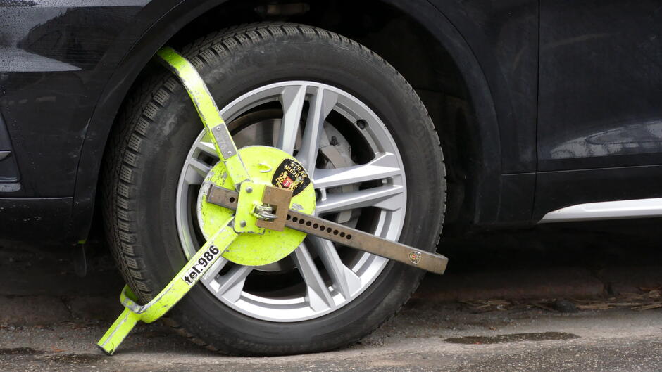 Na zdjęciu widać koło samochodu z założoną blokadą na koło. Blokada jest metalowa, w kolorze jaskrawożółtym, z widocznym numerem 986 i mechanizmem zamykającym w centralnej części. Koło ma aluminiową felgę o nowoczesnym, wieloramiennym wzorze. Samochód jest ciemnego koloru, prawdopodobnie czarny lub granatowy, a pojazd stoi na asfaltowej nawierzchni. Blokada uniemożliwia poruszanie się pojazdu, co sugeruje, że został on zaparkowany w miejscu niedozwolonym lub w związku z innym naruszeniem przepisów.