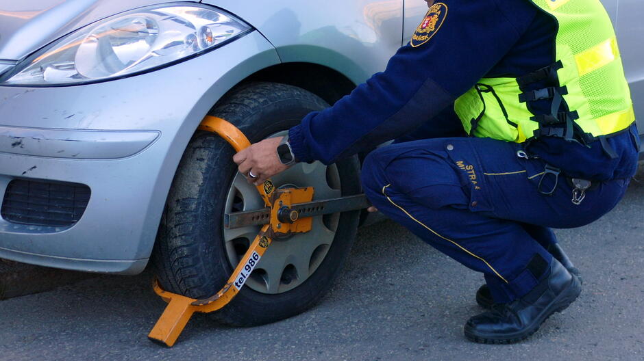 Na zdjęciu widać funkcjonariusza Straży Miejskiej zakładającego blokadę na koło samochodu osobowego. Pojazd jest koloru srebrnego, a blokada ma intensywnie pomarańczowy kolor z widocznym numerem identyfikacyjnym 986. Funkcjonariusz ubrany jest w granatowy mundur z odblaskową kamizelką w kolorze żółtym, a na pasie ma przypięte wyposażenie służbowe. Scena odbywa się na utwardzonej nawierzchni, prawdopodobnie przy drodze lub parkingu. Blokada jest montowana na przednim lewym kole pojazdu, co wskazuje na interwencję związaną z nieprawidłowym parkowaniem lub innym naruszeniem przepisów.