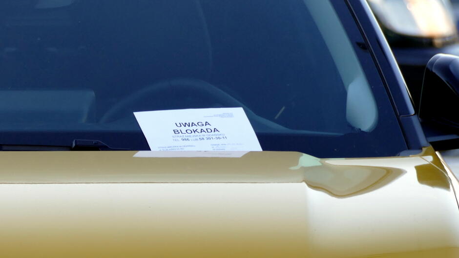 Na zdjęciu widać przednią szybę samochodu w kolorze złotym, na której umieszczona jest kartka z informacją „UWAGA BLOKADA”. Na kartce widoczny jest również numer telefonu do kontaktu w sprawie zdjęcia blokady. Pojazd stoi na zewnątrz, prawdopodobnie na parkingu, a w tle widać fragment innego samochodu. Kartka jest umieszczona na środku dolnej części szyby, co wskazuje, że pojazd został unieruchomiony przez służby porządkowe w związku z naruszeniem przepisów.