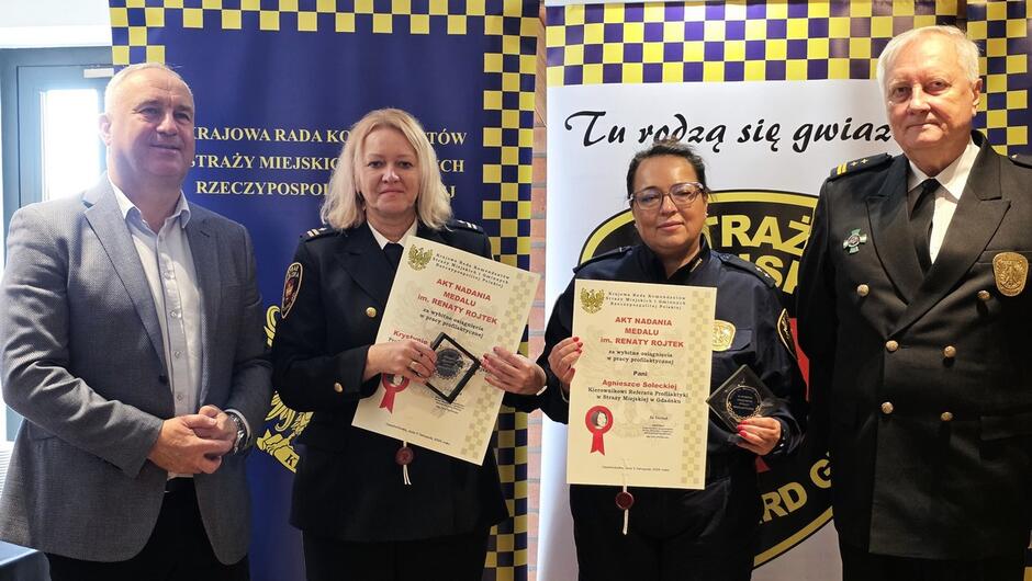 Zdjęcie grupowe uczestników wydarzenia.
Elementy:
Grupa kilkunastu osób ustawiona przed budynkiem z cegły i jasnej elewacji.
W centrum widoczny pomnik lub popiersie na cokole z napisem „RENATA ROJTEK”.
Większość osób w mundurach straży miejskiej, kilka w garniturach.
Tło: budynek z oknami, tablicą informacyjną i drzwiami wejściowymi.