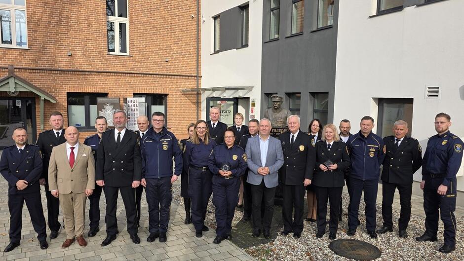 Wręczenie odznaczeń lub wyróżnień.
Elementy:
Cztery osoby stojące w rzędzie.
Dwie osoby w mundurach trzymają duże dyplomy z napisem „AKT NADANIA MEDALU im. RENATY ROJTEK” oraz ozdobne statuetki.
W tle te same banery co na pierwszym zdjęciu.
Widoczny tekst na dyplomach:
„AKT NADANIA MEDALU im. RENATY ROJTEK w uznaniu wybitnych osiągnięć w pracy samorządowej”