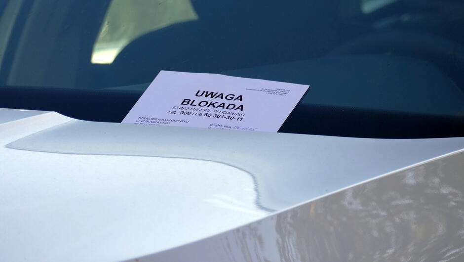 Na zdjęciu widać przednią szybę białego samochodu, na której umieszczona jest kartka z dużym, pogrubionym napisem „UWAGA BLOKADA”. Pod nagłówkiem znajduje się informacja o konieczności kontaktu z Strażą Miejską w Gdańsku oraz numery telefonów do dyżurnego. Kartka jest umieszczona za wycieraczką, co wskazuje na interwencję służb porządkowych w związku z nieprawidłowym parkowaniem. Tło zdjęcia jest rozmyte, skupiając uwagę na komunikacie.