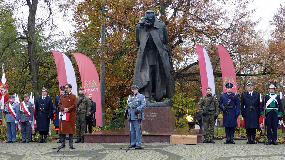 Na zdjęciu widoczna jest uroczystość patriotyczna przy pomniku marszałka Józefa Piłsudskiego. Centralnie znajduje się monumentalny pomnik na ciemnoczerwonym cokole, otoczony przez uczestników wydarzenia. Po obu stronach pomnika stoją osoby w mundurach wojskowych i historycznych, a także przedstawiciele służb mundurowych w galowych strojach. W tle widoczne są czerwone banery z herbem Gdańska oraz biało-czerwone flagi narodowe. Sceneria jest jesienna – drzewa w tle mają liście w odcieniach pomarańczu i brązu, a brukowany plac podkreśla oficjalny charakter miejsca. Na pierwszym planie, przed pomnikiem, ustawione są mikrofony, co sugeruje, że trwa część oficjalnych przemówień.
