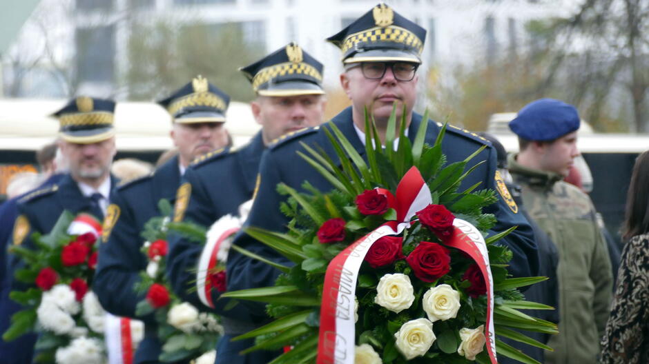 Na zdjęciu widać funkcjonariuszy Straży Miejskiej w galowych mundurach, stojących w szeregu i trzymających okazałe wieńce z biało-czerwonymi szarfami. Wieńce są udekorowane czerwonymi i białymi różami oraz zielonymi liśćmi, a na szarfach widnieje napis „Straż Miejska Gdańsk” oraz „Prezydent Miasta Gdańska”. W tle widoczne są inne osoby uczestniczące w uroczystości oraz fragmenty zabudowy miejskiej. Sceneria sugeruje oficjalne wydarzenie patriotyczne związane z obchodami Święta Niepodległości.