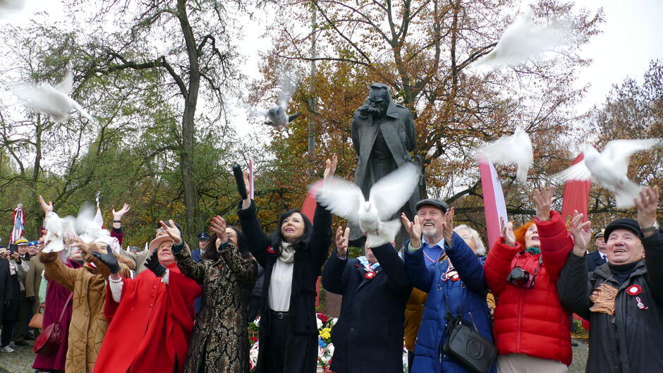 Na zdjęciu widać uroczysty moment wypuszczania białych gołębi w powietrze jako symbol pokoju i wolności. Na pierwszym planie stoją uczestnicy wydarzenia z uniesionymi rękami, wypuszczając ptaki. W tle znajduje się pomnik marszałka Józefa Piłsudskiego, otoczony czerwonymi banerami i biało-czerwonymi flagami. Sceneria jest jesienna – drzewa mają liście w odcieniach pomarańczu i brązu, a brukowany plac podkreśla oficjalny charakter uroczystości.
