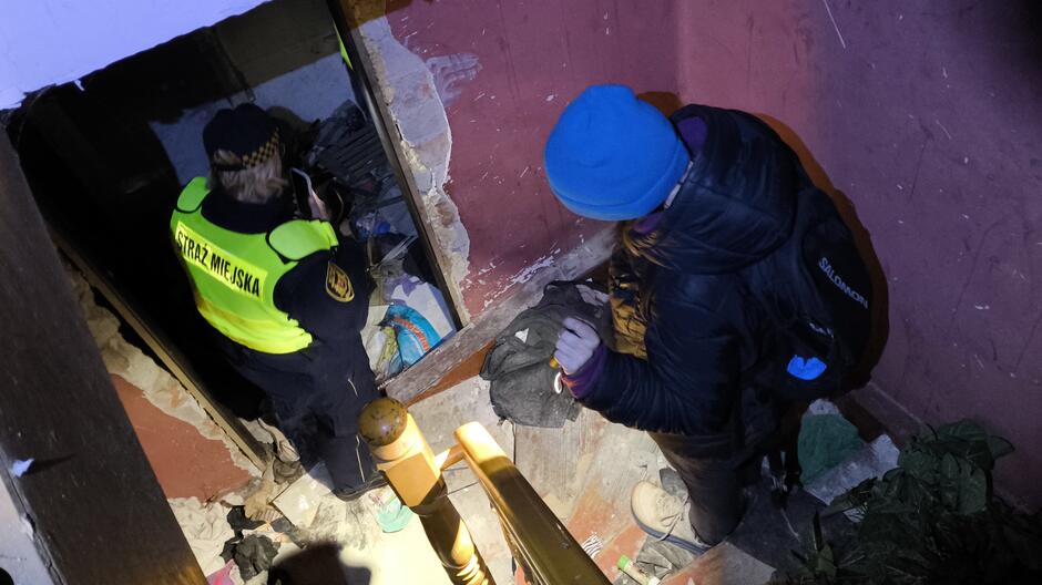 Na zdjęciu widać wnętrze zniszczonego, opuszczonego budynku. W centrum kadru znajduje się drewniana klatka schodowa z poręczą, na której pada światło latarki. Po prawej stronie stoi osoba w ciemnej kurtce i niebieskiej czapce, z plecakiem, trzymająca w dłoni latarkę. Po lewej stronie, w wejściu do małego pomieszczenia, znajduje się funkcjonariusz Straży Miejskiej w charakterystycznej żółto-czarnej kamizelce z napisem „STRAŻ MIEJSKA” na plecach. W tle widoczne są zniszczone ściany, odpadający tynk oraz porozrzucane rzeczy – ubrania, torby i śmieci, co wskazuje na miejsce wykorzystywane przez osoby w kryzysie bezdomności.