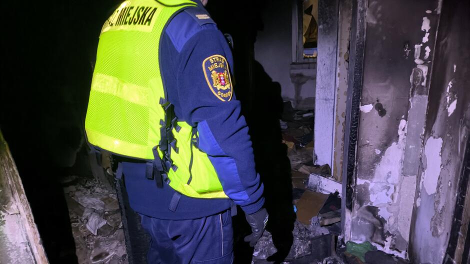 Na zdjęciu widać funkcjonariusza Straży Miejskiej ubranego w granatowy mundur i jaskrawożółtą kamizelkę z napisem „STRAŻ MIEJSKA” na plecach. Osoba stoi w zniszczonym, opuszczonym pomieszczeniu, oświetlonym światłem latarki. Ściany są mocno zabrudzone i nadpalone, z widocznymi śladami ognia i odpadającym tynkiem. Na podłodze leżą porozrzucane przedmioty – fragmenty kartonów, odzież i śmieci, co wskazuje na miejsce wykorzystywane przez osoby w kryzysie bezdomności. W tle widać kolejne zniszczone pomieszczenie.