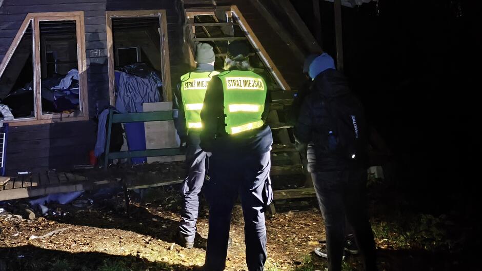 Na zdjęciu widać dwóch funkcjonariuszy Straży Miejskiej w jaskrawożółtych kamizelkach z napisem „STRAŻ MIEJSKA” na plecach oraz osobę towarzyszącą w ciemnej kurtce i czapce. Grupa stoi przed drewnianym budynkiem, który wygląda na opuszczony lub zniszczony – widać brak szyb w oknach, porozrzucane rzeczy wewnątrz oraz prowizoryczne wejście. Scena jest oświetlona światłem latarki, co sugeruje kontrolę prowadzoną po zmroku. W tle widoczne są schody prowadzące na górę budynku.