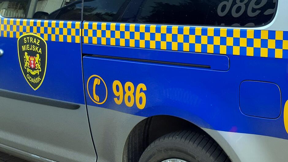 Na zdjęciu widoczny jest fragment pojazdu Straży Miejskiej w Gdańsku. Samochód ma charakterystyczne oznakowanie: niebieski pas z żółtą szachownicą biegnącą wzdłuż górnej części drzwi, herb Gdańska z napisem „STRAŻ MIEJSKA GDAŃSK” oraz numer alarmowy 986 w kolorze żółtym obok symbolu telefonu. Widać również część tylnego koła i srebrny lakier nadwozia. Zdjęcie zostało wykonane z bliska, skupiając się na bocznej części pojazdu.