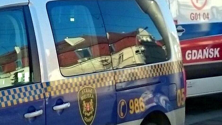 Na zdjęciu widać fragment pojazdu Straży Miejskiej w Gdańsku oraz częściowo widoczny ambulans. Samochód straży jest oznakowany charakterystycznym niebieskim pasem z żółtą szachownicą, herbem Gdańska z napisem „STRAŻ MIEJSKA GDAŃSK” oraz numerem alarmowym 986 obok symbolu telefonu. W tle znajduje się biało-czerwony ambulans z napisem „GDAŃSK” i oznaczeniem „G01 04”. Na powierzchni pojazdów odbijają się budynki i linie napowietrzne, co sugeruje, że zdjęcie wykonano na ulicy w ciągu dnia.