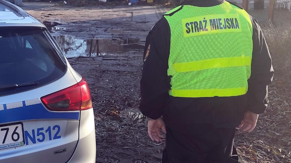 Na pierwszym planie widać funkcjonariusza Straży Miejskiej ubranego w czarny mundur z jaskrawożółtą kamizelką odblaskową, na której widnieje napis „STRAŻ MIEJSKA”. Obok niego znajduje się fragment radiowozu policyjnego z widoczną częścią tylnej klapy, lampy oraz oznaczeniem N512 na tablicy rejestracyjnej. Tło zdjęcia przedstawia nieutwardzoną, błotnistą nawierzchnię z kałużami oraz elementami zabudowy w oddali. Scena wygląda na miejsce interwencji lub patrolu w terenie.